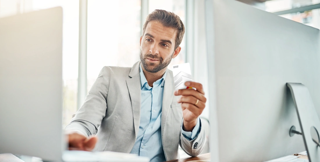 a man in a grey suit jacket smiles as he holds a payment card and looks at his laptop